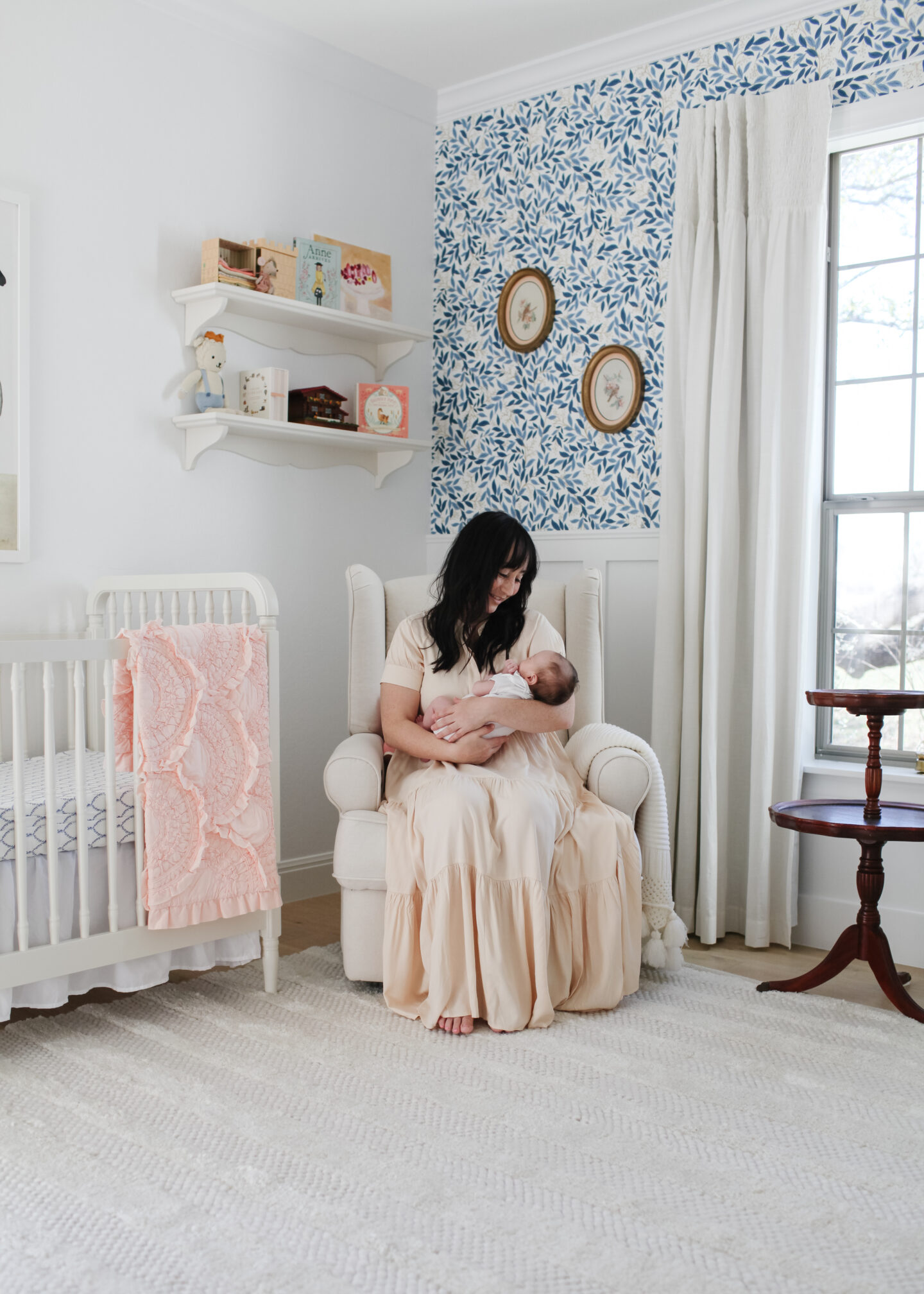 Baby girl nursery with blue and white wallpaper, floating shelves, crib, curtains, vintage pie crust table and rug